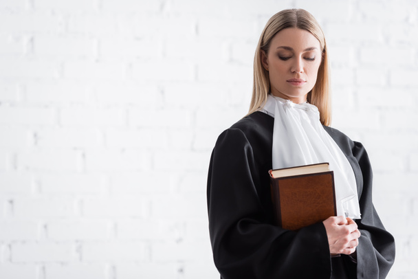procureur blonde en manteau noir debout avec livre près du mur blanc - Photo, image