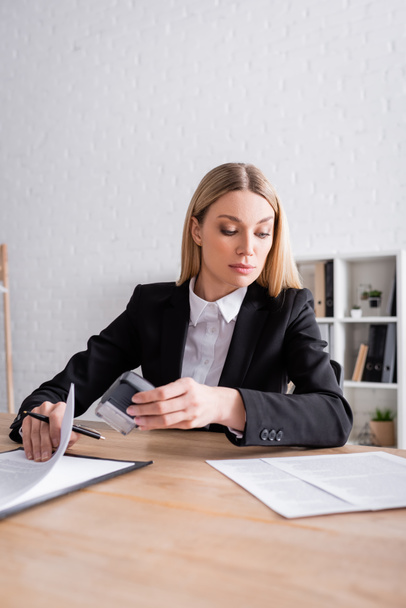 blonde lawyer in formal wear holding stamper and pen while looking at documents - Фото, изображение