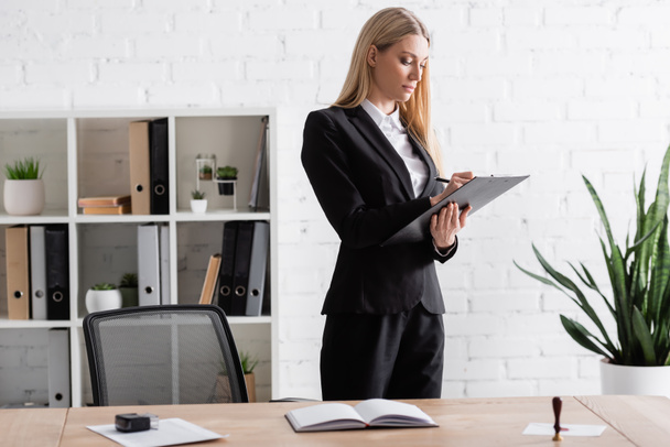 blonde lawyer writing on clipboard while standing in office - Photo, Image