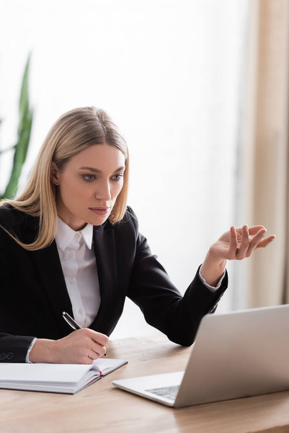 blonde notary gesturing near laptop while holding pen near notebook - Photo, Image