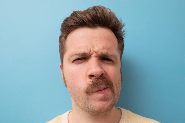 Young man with funny mustache having doubts and with confuse face expression on isolated blue background - Photo, Image