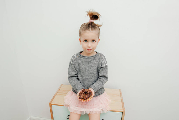 Cute little girl with funny hairstyle is sitting on the commode with chocolate donut - Φωτογραφία, εικόνα