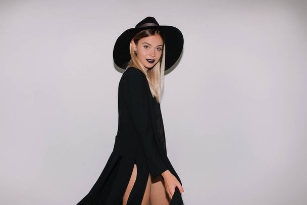 Portrait of young lady with holiday make up is standing over isolated background. Gorgeous blonde is wearing dark dress and black hat against white wall - Fotoğraf, Görsel