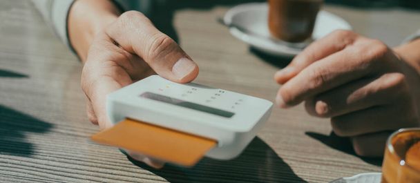 Крупный план молодого человека, сидящего за столом тротуарного кафе, оплачивающего счет своей кредитной картой, вставленной в беспроводной платежный терминал, в панорамном формате для использования в качестве веб-баннера или заголовка - Фото, изображение