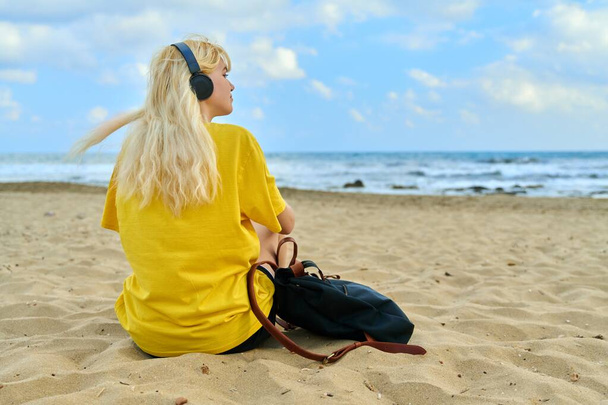 Modern teen female blonde in headphones with a backpack sitting on beach - Foto, Imagen