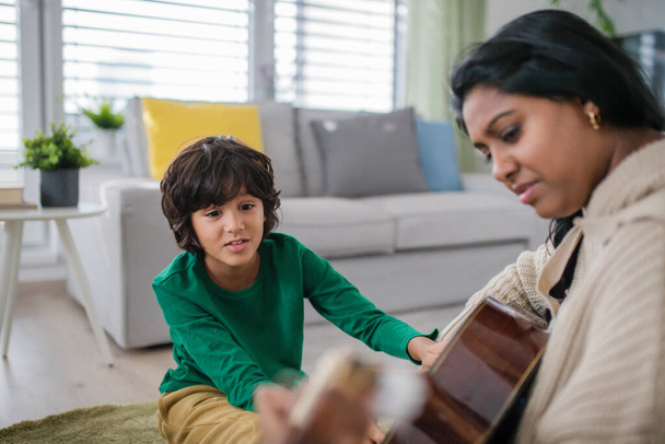 Little multiracial boy learning to play the guitar with his mother at home. - Photo, Image