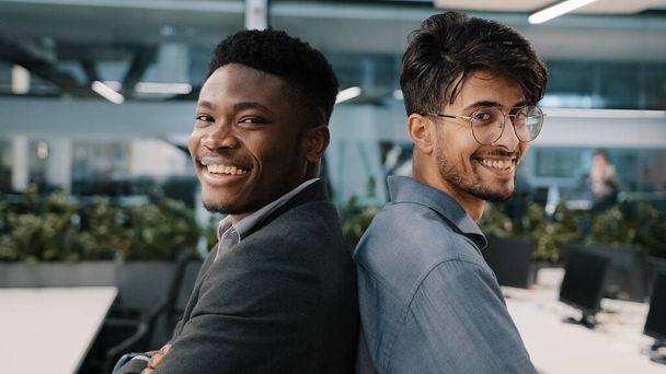 Portré két multinacionális férfi kollégák férfiak Afrikai arab indiai menedzser pózol irodai cég áll vissza egymással partnerség nézi kamera mosolygós üzleti partnerek alkalmazottak - Fotó, kép