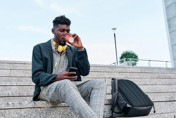 Εξωτερική πορτρέτο ενός μοντέρνου Αφροαμερικανού άνδρα πίνοντας καφέ σε ένα χάρτινο κύπελλο και ψάχνοντας για νέα ενδιαφέροντα μέρη σε ιστοσελίδες χρησιμοποιώντας μια online εφαρμογή στο κινητό τηλέφωνο. - Φωτογραφία, εικόνα