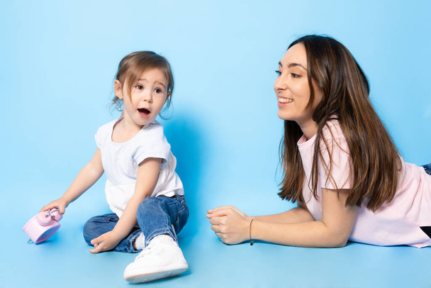 Bild einer glücklichen jungen Frau Mutter mit ihrem kleinen Mädchen Kind Tochter sitzt isoliert über blauem Hintergrund. - Foto, Bild