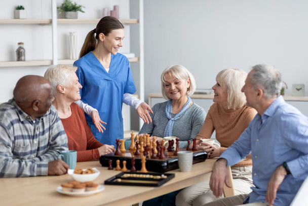 Friendly nurse talking to elderly people playing table games - Фото, изображение