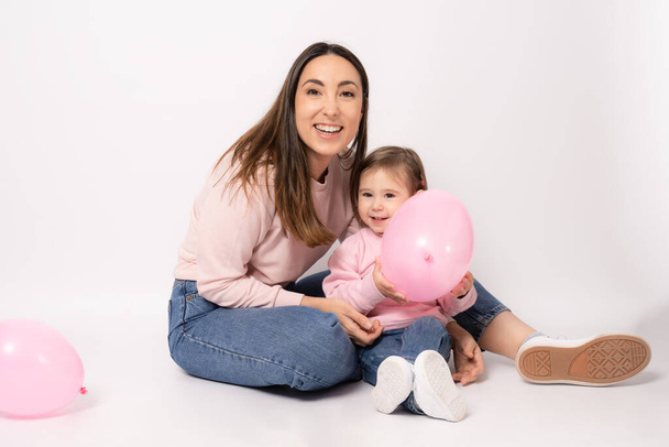 Glückliche Mutter mit kleiner Tochter sitzt auf dem Boden mit rosa Luftballons isoliert über weißem Hintergrund. - Foto, Bild