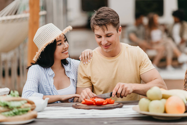 Młoda, zróżnicowana para gotująca razem w pobliżu kampera na świeżym powietrzu, krojąca warzywa na śniadanie, ciesząca się wycieczką - Zdjęcie, obraz