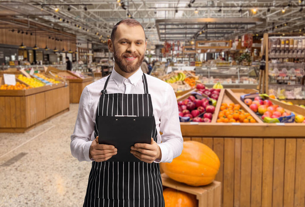 Male shop assistant in a grocery store holding a clipboard in front of fruits and vegetables - Photo, Image