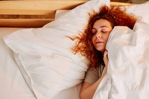 Una hermosa chica pelirroja está acostada en su cama y duerme apretada por la mañana justo antes de la alarma. - Foto, imagen