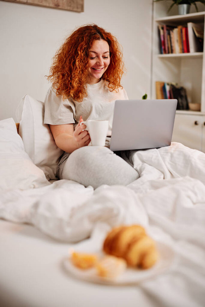 Una chica pelirroja se sienta en la cama por la mañana, bebiendo su café de la mañana y usando su computadora portátil. - Foto, Imagen
