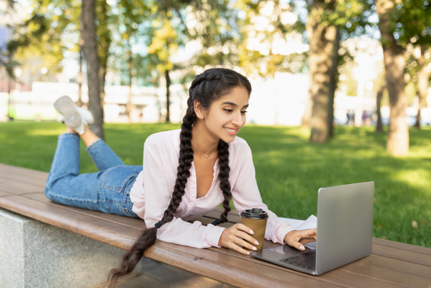 Descansa en el campus. Chica estudiante universitaria india feliz relajarse al aire libre con café y computadora portátil, acostado en el banco en el parque - Foto, imagen