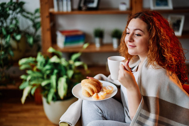 Une gingembre heureuse assise à la maison, tenant une bonne tasse chaude de café du matin et prenant le petit déjeuner. - Photo, image