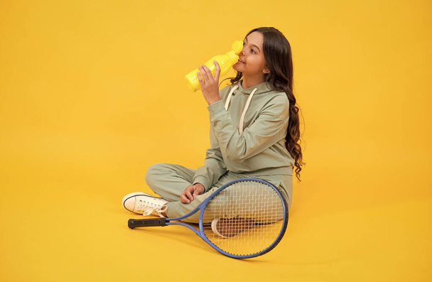 šťastný dospívající dívka držet badminton raketa a pít vodu z láhve na žlutém pozadí, relaxovat. - Fotografie, Obrázek