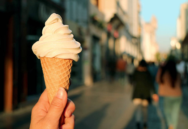 Крупный план Vanilla Soft Serve Ice Cream Cone in Hand with Blurry Walking Street in Backdrop - Фото, изображение