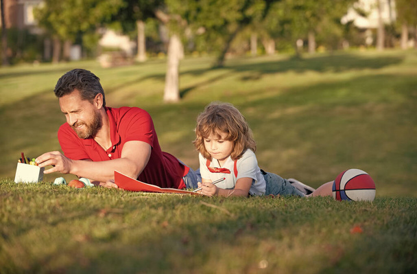 Feliz día de familia. Papá y el niño se relajan en el césped en el parque. niño mantenga aprender a dibujar. - Foto, imagen