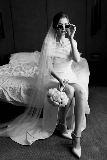 Stylowa panna młoda w sukni ślubnej pozująca w białych okularach siedząca na łóżku. Czarno-białe zdjęcie. Wysokiej jakości zdjęcie - Zdjęcie, obraz