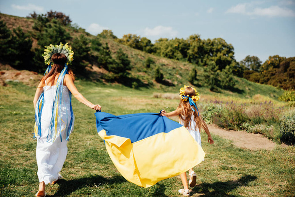 mother and daughter in traditional Ukrainian wreaths blue and yellow flag of Ukraine in field. - Фото, изображение