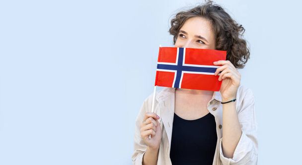 Young happy woman looking away peeking out from behind Norway flag while standing in front of gray background. - Foto, Imagem