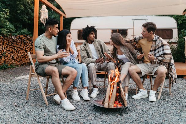 Diverse junge Freunde sitzen am Lagerfeuer und erzählen gruselige Geschichten neben Wohnmobil während der Zeltreise im Sommerurlaub - Foto, Bild
