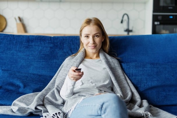 Zdjęcie szczęśliwej młodej kobiety patrzącej na kamerę trzymającą zdalnie sterowany program TV. Kobieta odpoczywa w weekend, siedzi na kanapie w salonie. Koncepcja rozrywki - Zdjęcie, obraz