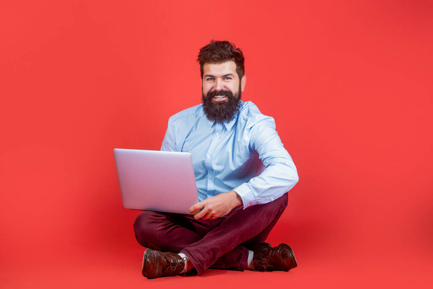 Pitelen kannettavaa tietokonetta. Hymyilevä komea parrakas mies työntekijä kannettava tietokone. Onnellinen nuori mies istuu lattialla kannettavan tietokoneen kanssa ja käyttää sitä punaisella taustalla - Valokuva, kuva