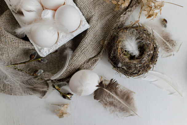 Simple rural Easter aesthetics. Natural eggs in tray, feathers, willow branches, nest on burlap on rustic white wooden table. Happy Easter. Easter rustic still life. - Photo, Image