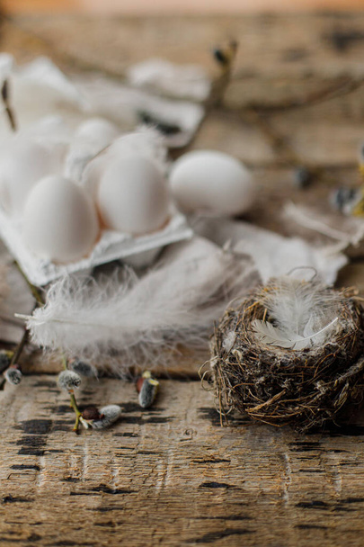 Stylish Easter rustic still life. Easter natural eggs in tray, feathers, willow branches, nest, linen cloth on aged wooden table. Happy Easter! Simple rural aesthetics - Photo, Image