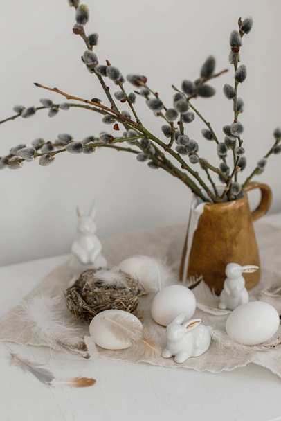 Easter rustic still life. Natural eggs, bunny figurines, feathers, nest, willow branches in vase on aged wooden table. Simple stylish easter decoration on table. Happy Easter - Photo, Image
