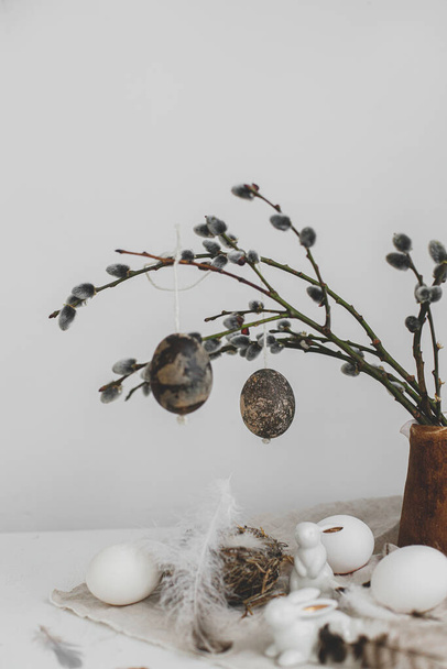 Easter rustic still life. Natural eggs, bunny figurines, feathers, nest, willow branches in vase on aged wooden table. Simple stylish easter decoration on table. Happy Easter - Photo, Image