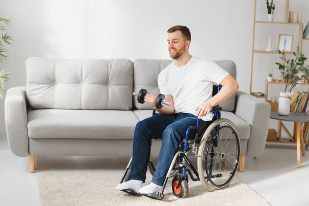 sportsman sitting in wheelchair and outstretching arms with dumbbells during rehabilitation exercise in modern medical center. Man sitting on wheelchair at home. - Foto, imagen