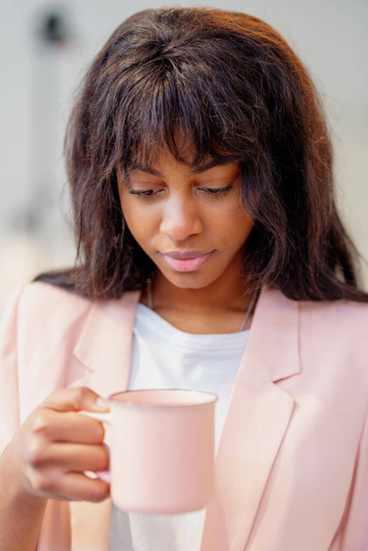 jong Afrikaans meisje elegant gekleed in een koraal gekleurde pak met een koraal gekleurde koffiekop tijdens haar werkdag in een kantoor. - Foto, afbeelding