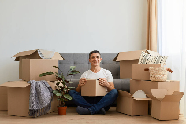 Indoor shot of attractive smiling delighted man wearing white t shirt sitting on floor near sofa surrounded with cardboard boxes, holding carton parcel, relocating to a new flat. - Fotoğraf, Görsel