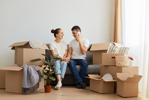 Horizontal shot of optimistic young couple sitting on sofa surrounded with carton boxes, family during moving in, man with happy smile talking via mobile phone. - Foto, Imagen