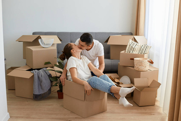 Portrait of happy romantic couple having fun while moving into new house, attractive woman sitting in cardboard box while man kissing her with love na gentle. - Valokuva, kuva