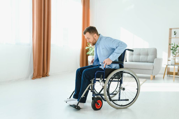 Disabled young guy trying to get up from wheelchair at home, copy space. - Foto, Imagen