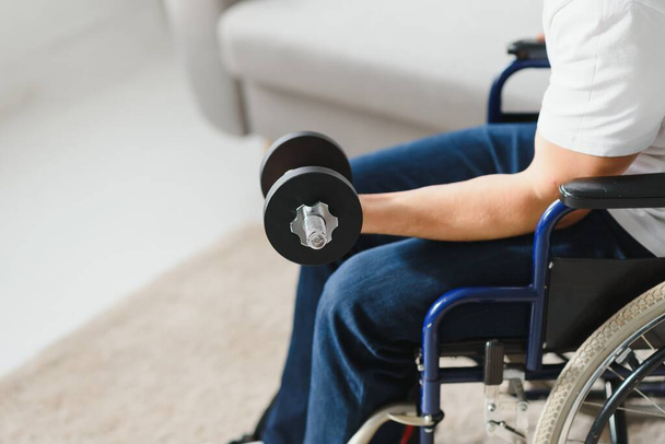 deportista sentado en silla de ruedas y brazos extendidos con pesas durante el ejercicio de rehabilitación en el centro médico moderno. Hombre sentado en silla de ruedas en casa. - Foto, imagen