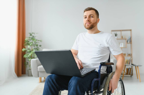 Portrait of smiling disabled male sitting in wheelchair and working on laptop from home. Young worker with special needs. Freelancer and people with disabilities concept. - Fotoğraf, Görsel