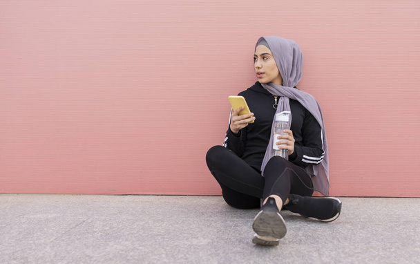 Молодая мусульманка пьет воду. Молодая мусульманка держит в руках бутылку воды и мобильный телефон. сидя на земле, отдыхая - концепция арабской спортсменки -. - Фото, изображение