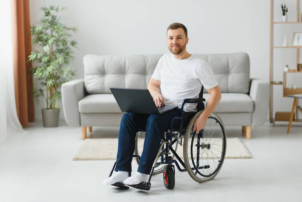 車椅子に座り、自宅からノートパソコンで作業する障害者の笑顔の肖像. - 写真・画像