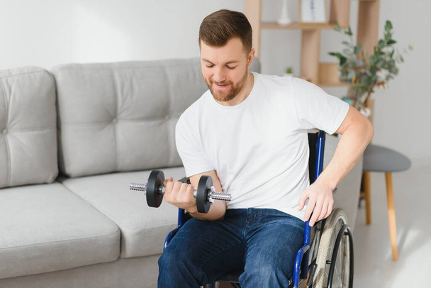 sportsman sitting in wheelchair and outstretching arms with dumbbells during rehabilitation exercise in modern medical center. Man sitting on wheelchair at home. - Foto, Bild