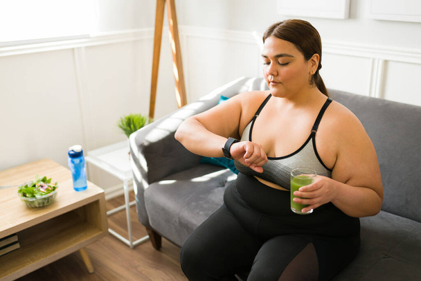 Ernährung und grüner Saft. Junge fettleibige Frau schaut sich die Gesundheits-App auf ihrer Smartwatch an und zählt Kalorien, um abzunehmen - Foto, Bild