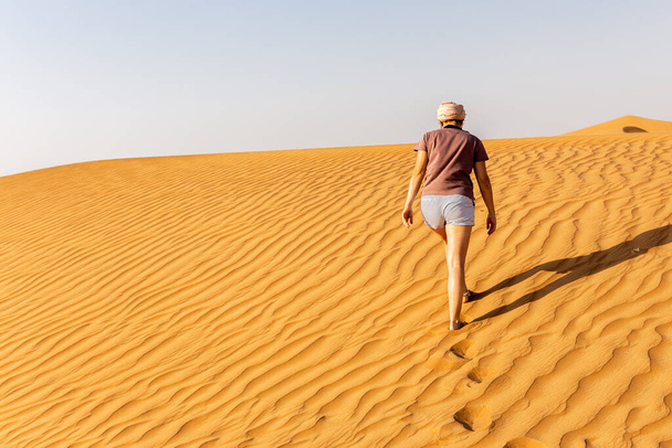 Young caucasian woman in shorts and t-shirt walking alone on a sand dune towards the arabian desert, footprints and ripples in the sand, United Arab Emirates. - Foto, Imagen