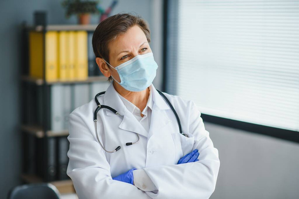 Older female doctor wearing face mask and white medical coat standing in hospital. Portrait - Foto, Imagen