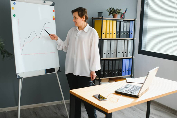 Mature older businesswoman mentor coach training speaker give flip chart business presentation at company meeting, confident female executive manager teaching team consult clients at office workshop - 写真・画像