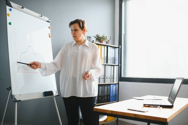 Smiling old mature female coach manager trainer pointing on whiteboard give business flip chart presentation, happy senior adult teacher mentor speaker presenter conduct conference workshop concept - 写真・画像